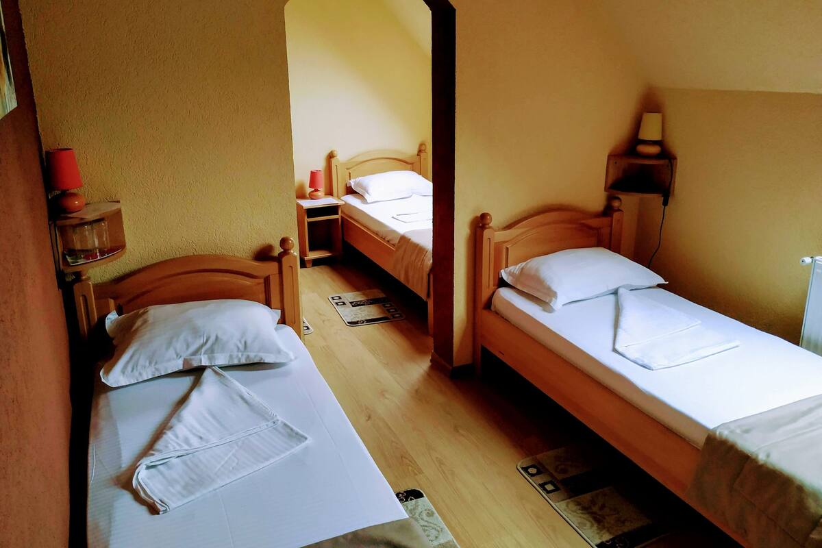 Quarto com duas camas de solteiro, cabeceiras de madeira, uma mesa de cabeceira com abajur e uma pequena prateleira com abajur vermelho e um recipiente de vidro.