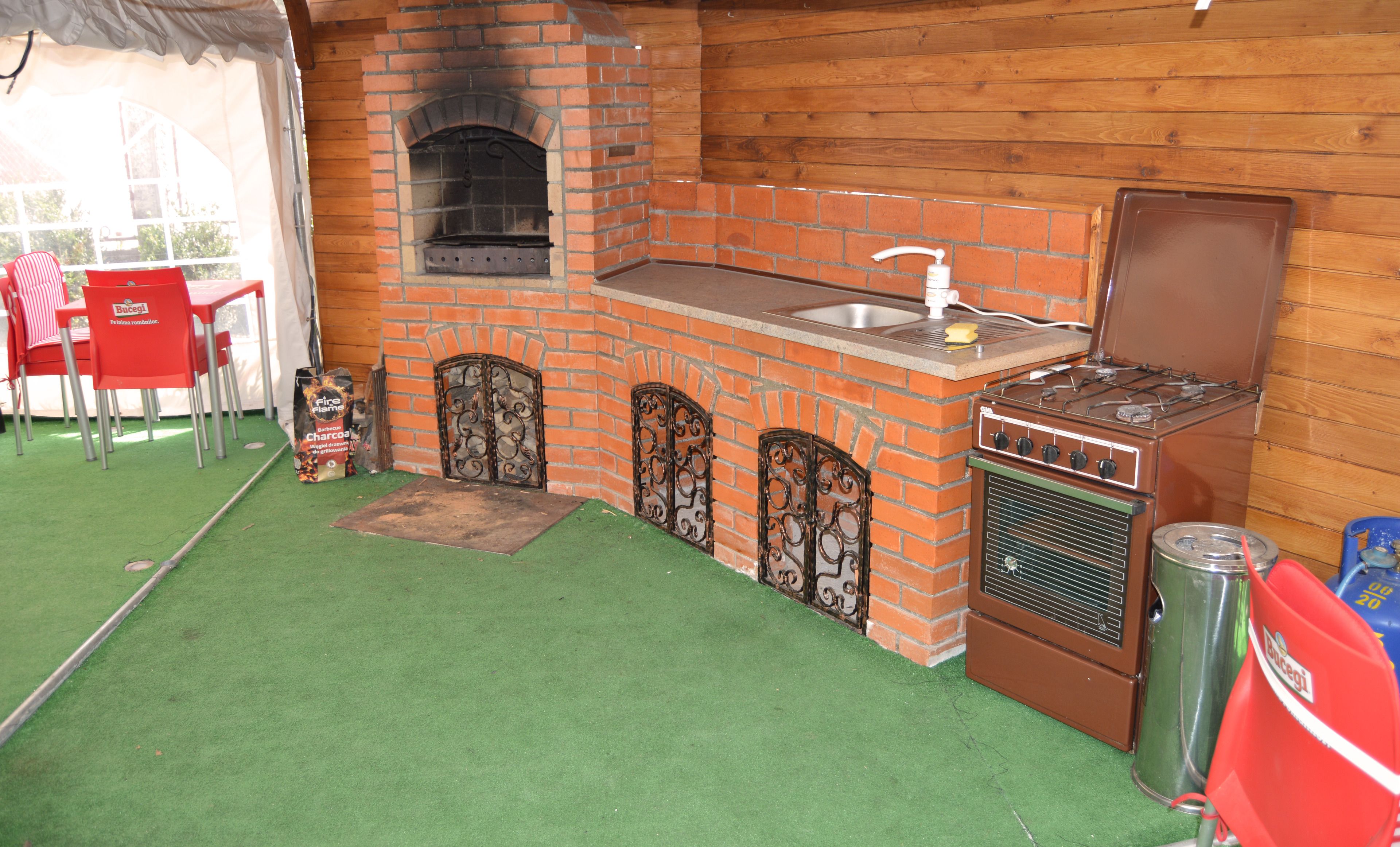 Een buitenkeuken met een bakoven, een spoelbak en een kookplaat. Rode stoelen en een tafel zijn zichtbaar.