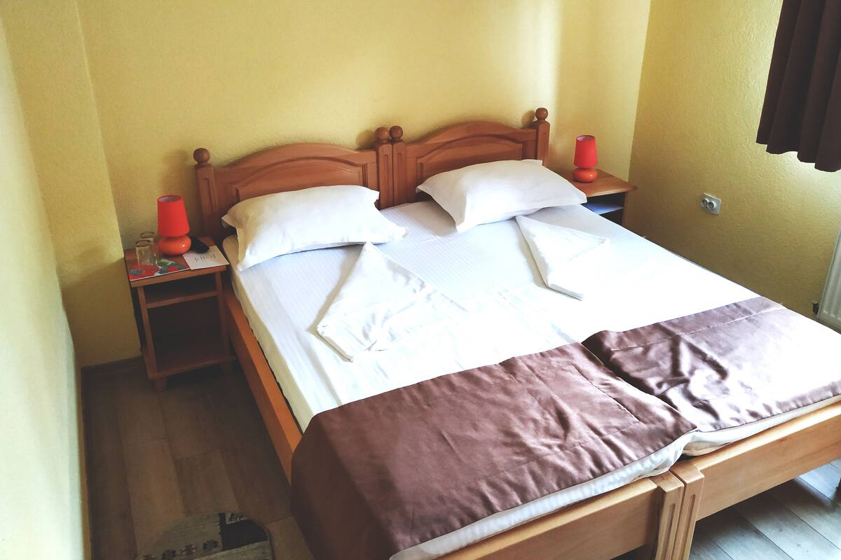 Cama bem arrumada com lençóis brancos e manta marrom, cabeceira de madeira e abajur vermelho sobre a mesa de cabeceira.