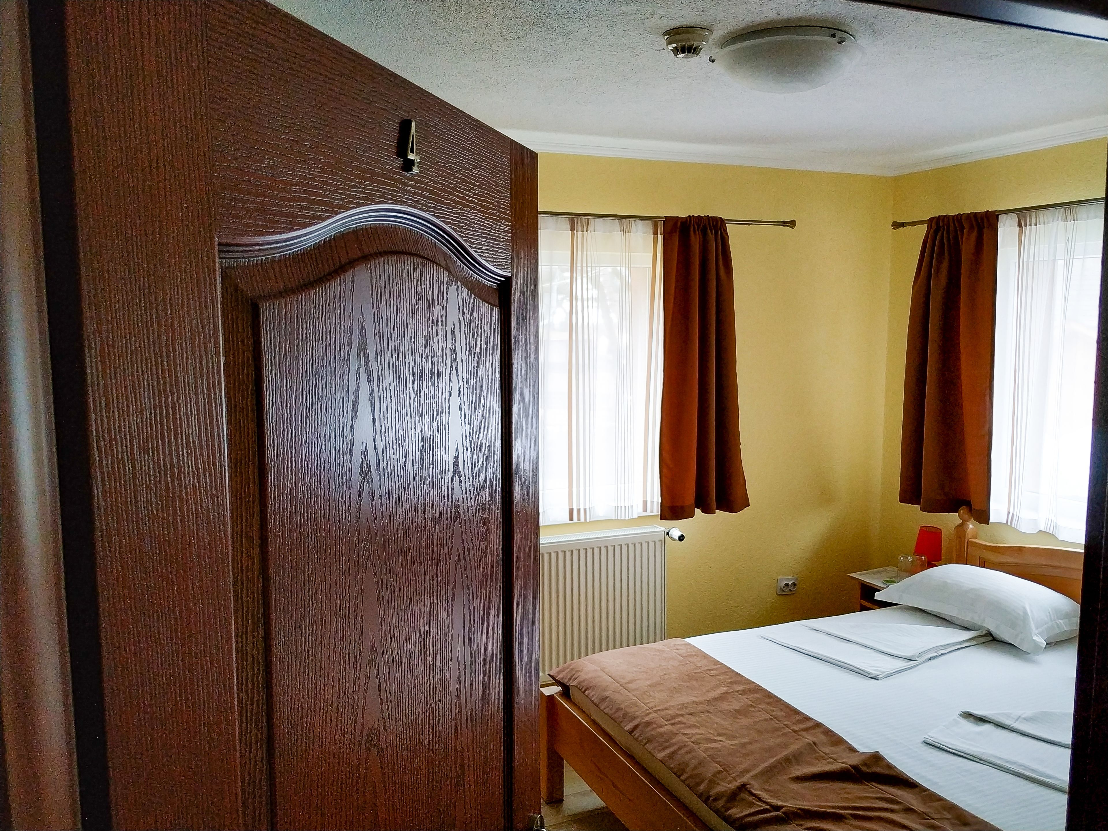 A bedroom with a wooden wardrobe, a bed with white linens, a bedside table with a lamp, and a window with curtains.