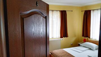 A bedroom with a wooden wardrobe, a bed with white linens, a bedside table with a lamp, and a window with curtains.