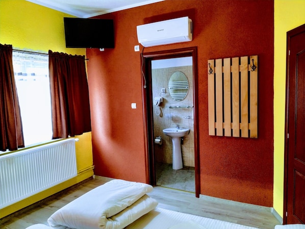 A hotel room with a bed, a window with curtains, a mounted TV, an air conditioning unit, a bathroom with a sink and mirror, and wooden slats on the wall. - Venesis House (Sighisoara)
