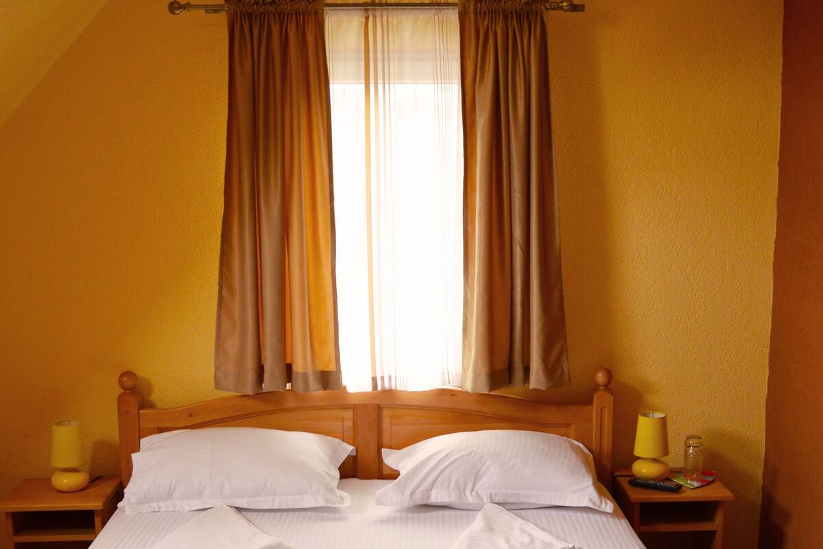 Cama bem arrumada em quarto com paredes amarelas, cabeceira de madeira e janela com cortinas. Duas mesas de cabeceira, com luminárias e um controle remoto em uma delas.