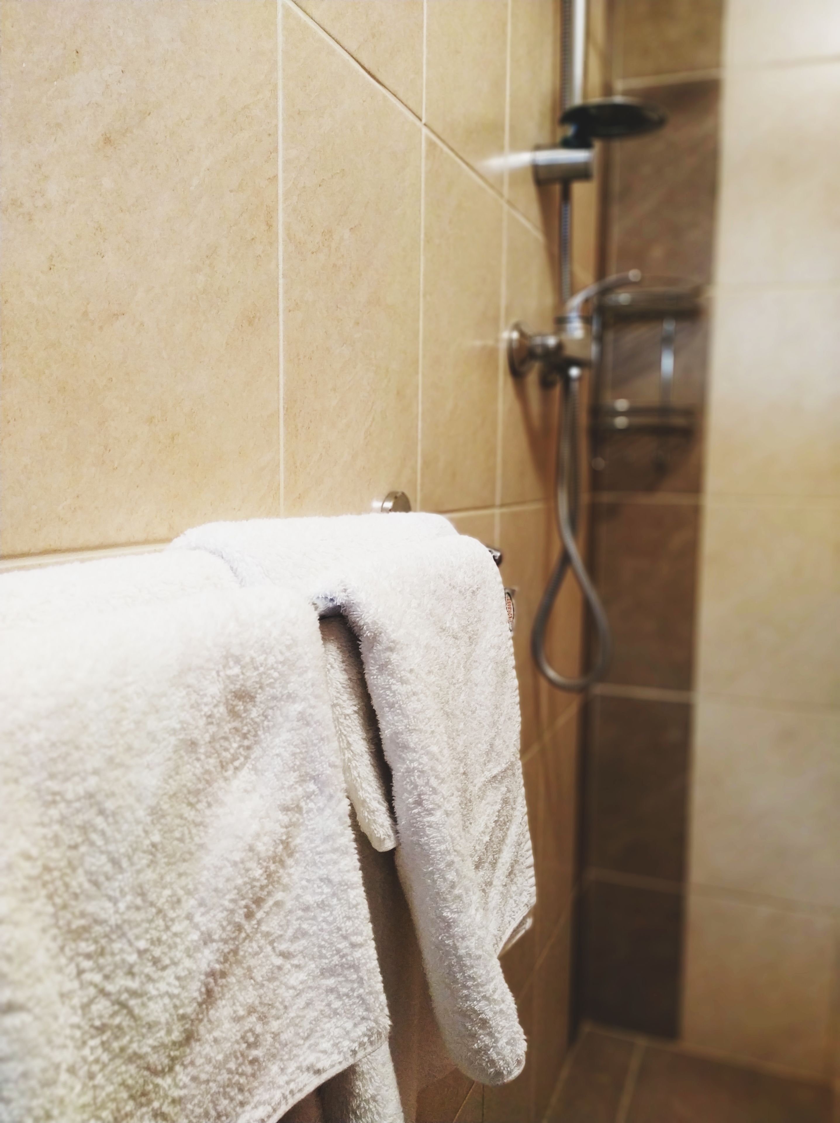 Un baño con baldosas color beige, una toalla blanca en un perchero y un cabezal de ducha.
