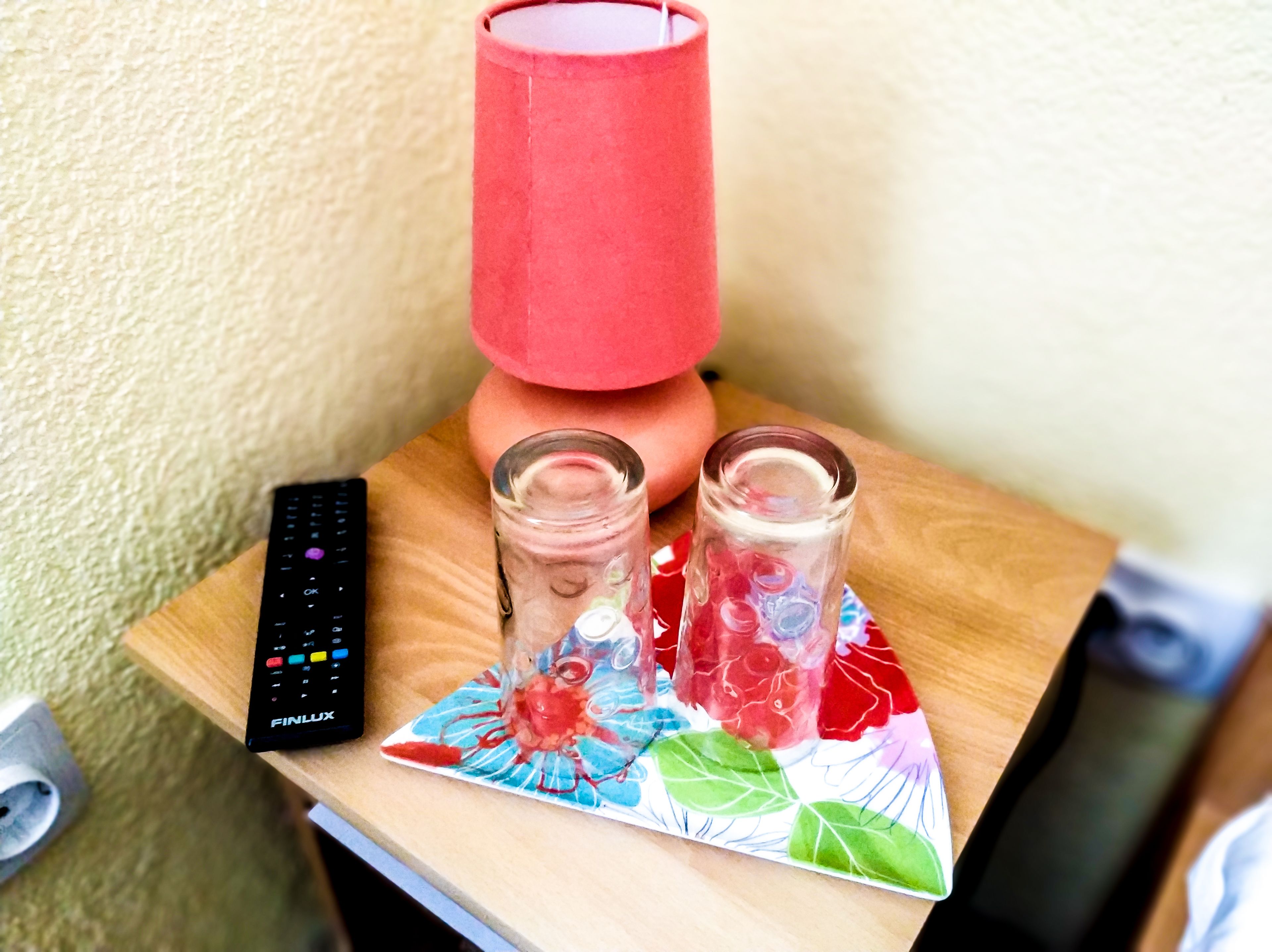 Een afstandsbediening, twee glaasjes, een rode lamp en een decoratieve onderzetter met bloemenprint op een houten tafel.