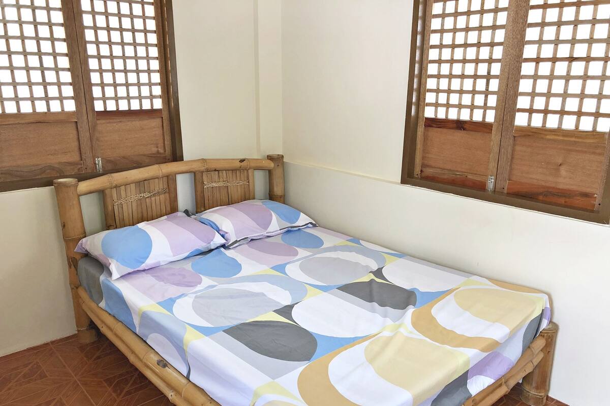 A bed with a patterned bedspread and pillows, a wooden headboard, and a window with a wooden frame.