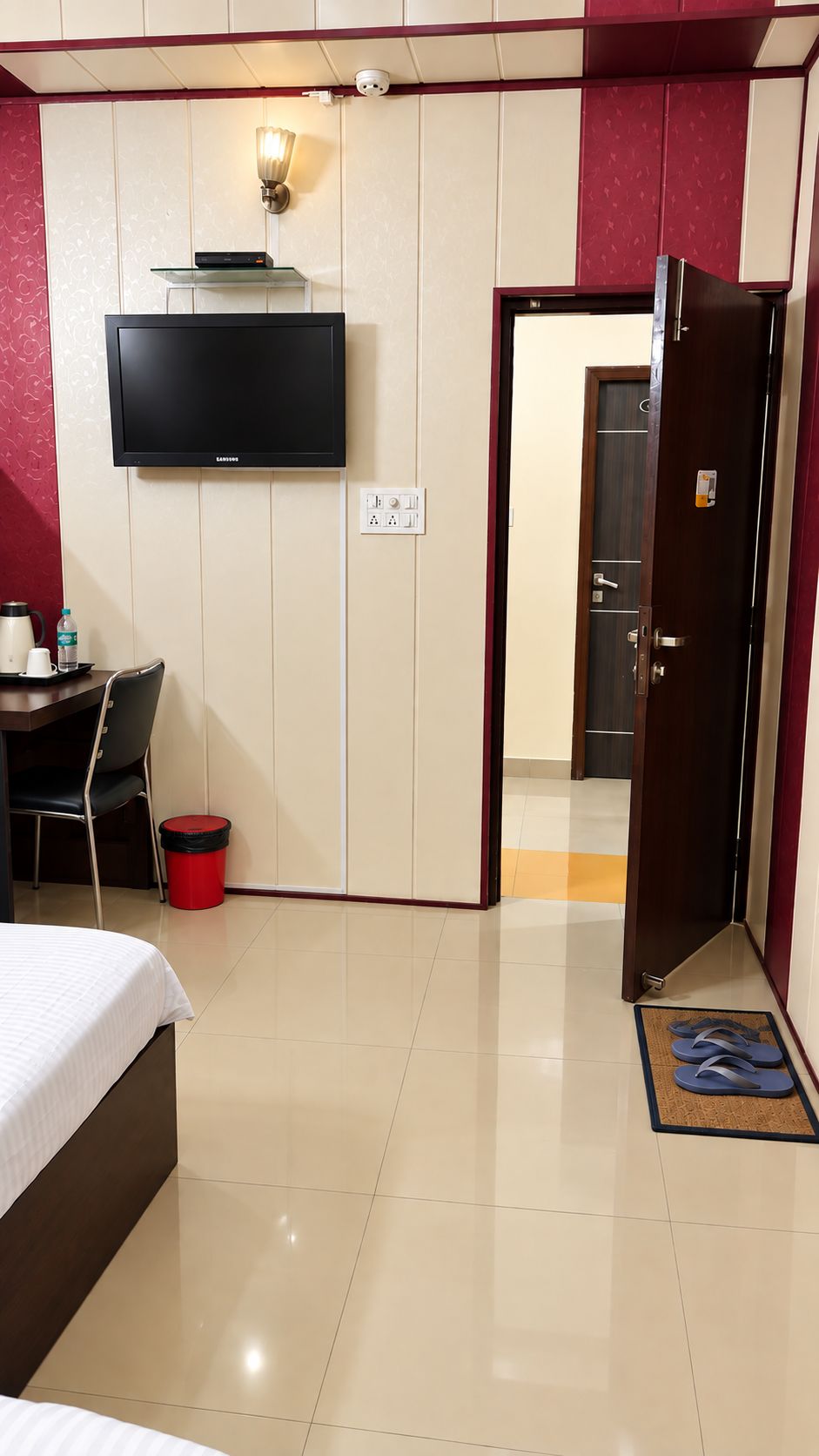 A hotel room with a bed, a desk with a chair, a television mounted on the wall, and a door leading to another room.