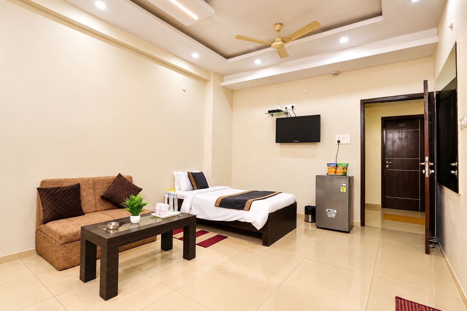 A hotel room with a sofa, a bed, a small table, a TV, and a minibar.