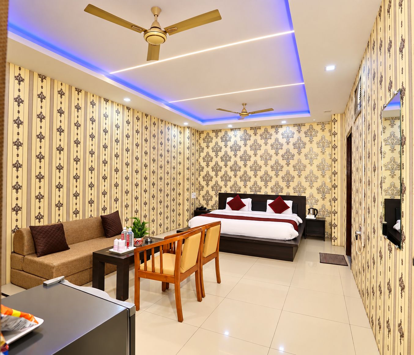 A hotel room with a bed, a sofa, a desk, and a chair. There are two ceiling fans and a TV mounted on the wall.