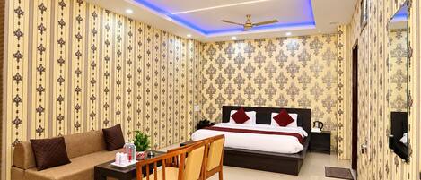 A hotel room with a bed, a sofa, a desk, and a chair. There are two ceiling fans and a TV mounted on the wall.