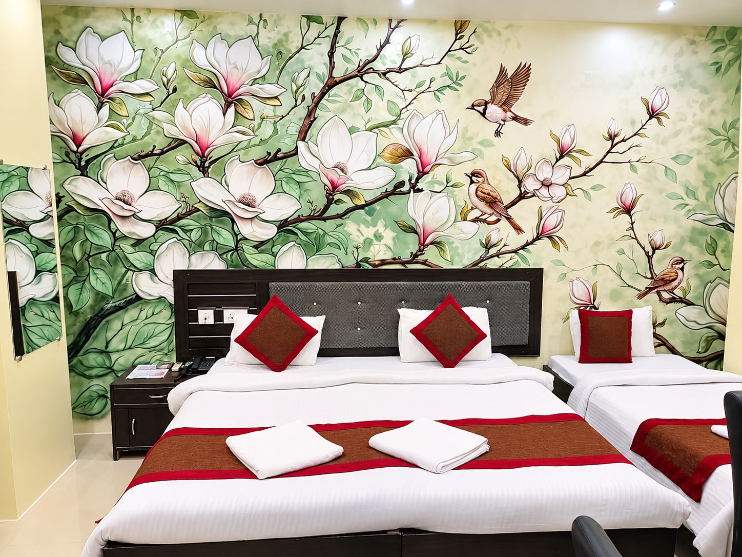 A hotel room with a large floral mural on the wall, a bed with white bedding and red accents, and a nightstand with a lamp.