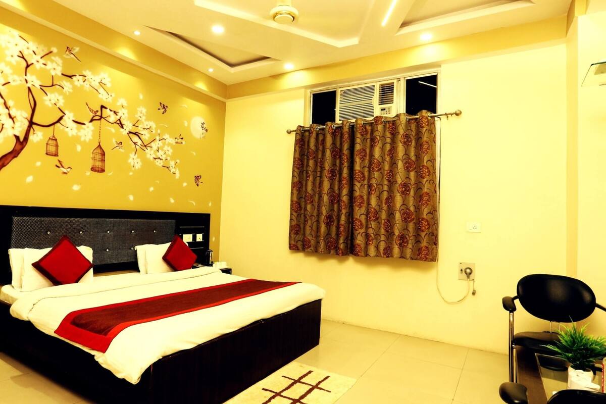 A hotel room with a bed, a nightstand, a desk with a chair, a window with curtains, and a wall with a floral mural.
