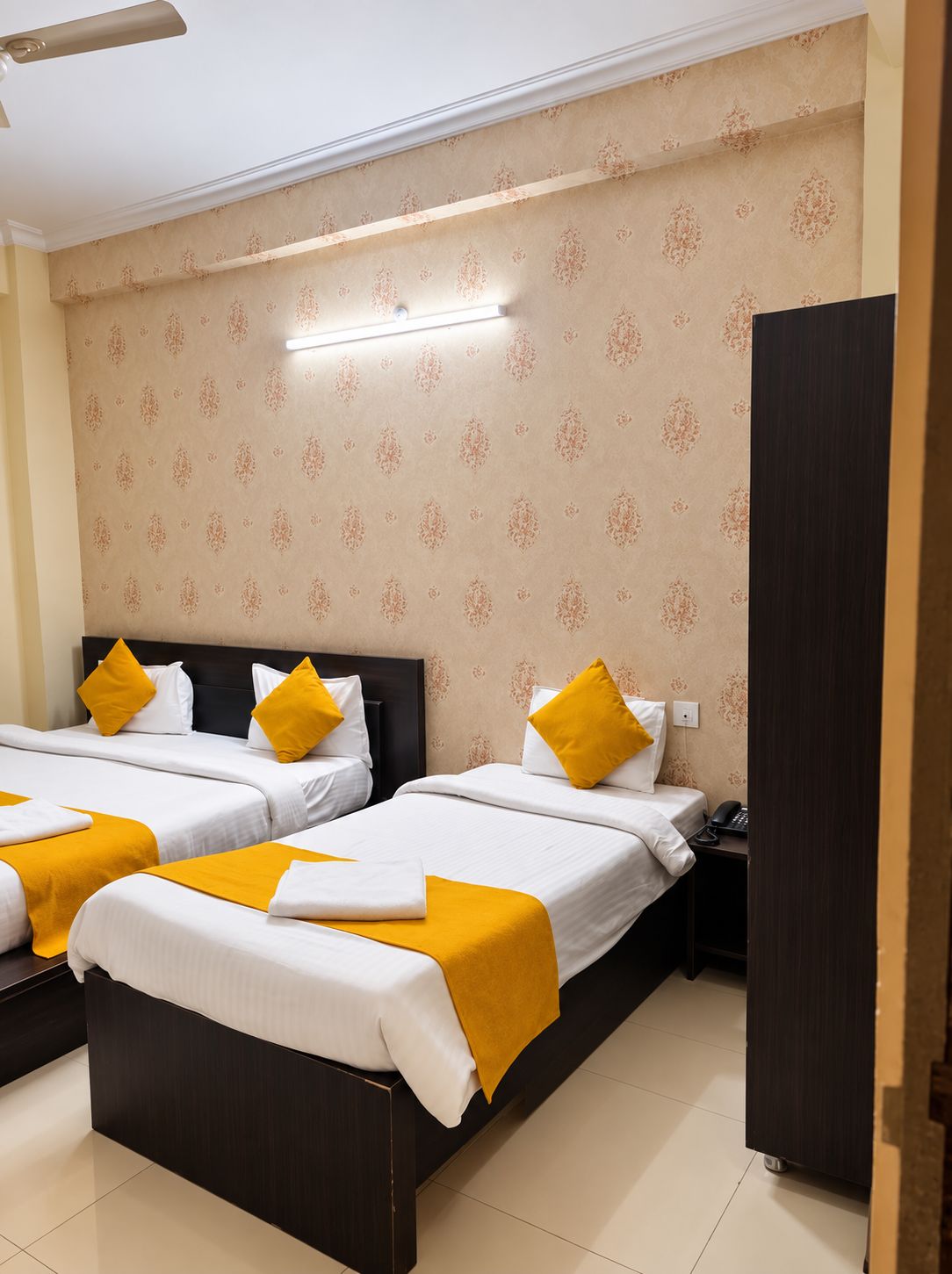 A hotel room with two beds, each with white bedding and yellow accents, a dark wooden headboard, a wardrobe, and a patterned wallpaper.