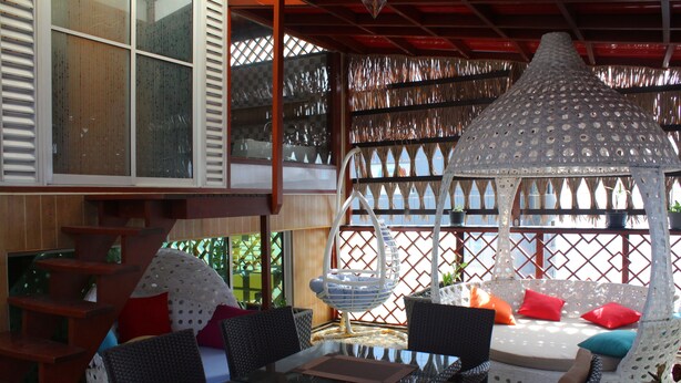A glass-top dining table with wicker chairs, a hanging light fixture, and a seating area with cushions and a decorative hanging basket. - Six In One (Malé)