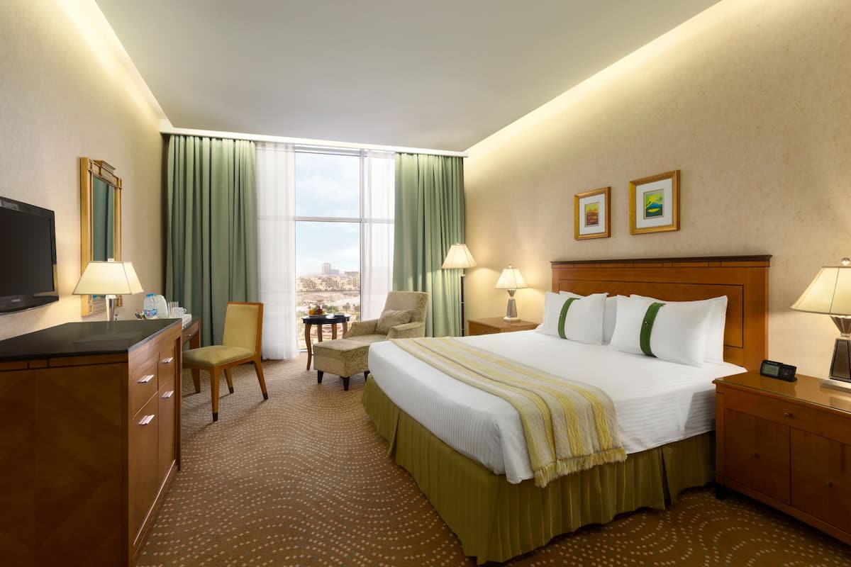 Habitación de hotel con una cama grande, un escritorio, una silla, un televisor y un ventanal con cortinas.