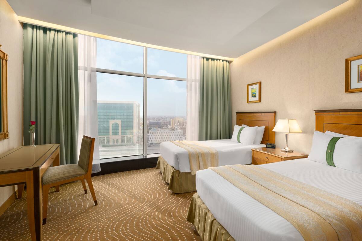 Habitación de hotel con dos camas, un escritorio, una silla, una lámpara y vistas a la ciudad a través de amplias ventanas.