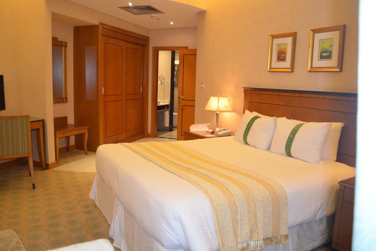 Habitación de hotel con una cama grande, armario de madera, un escritorio con lámpara y un baño visible a través de una puerta abierta.