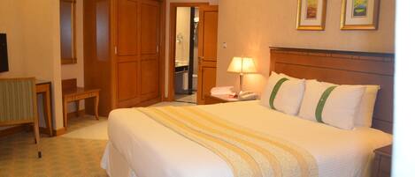 A hotel room with a large bed, wooden wardrobe, a desk with a lamp, and a bathroom visible through an open door.