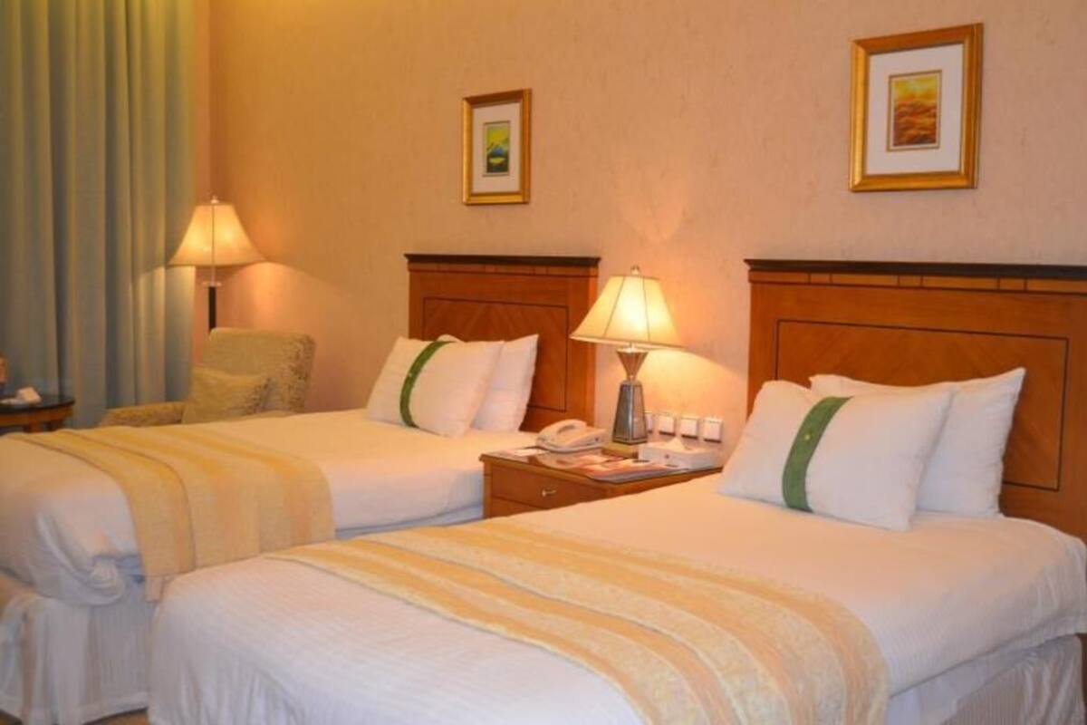 Habitación de hotel con dos camas, mesitas de noche, lámparas, teléfono y cuadros en la pared.