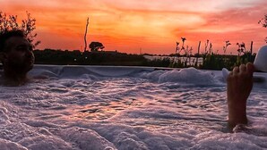 Un atardecer sobre un cuerpo de agua con olas, silueta de un árbol y una mano en el agua.
