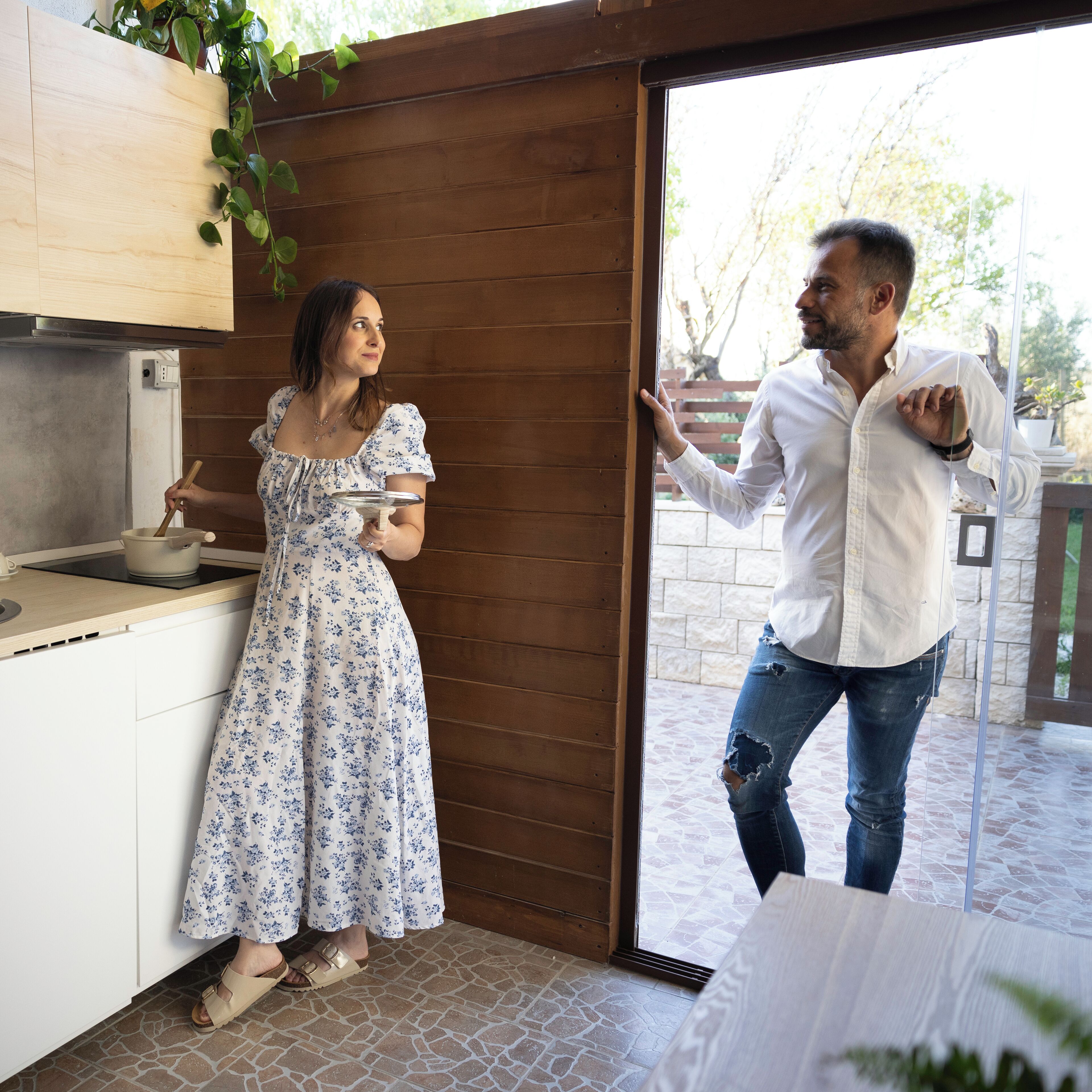 Une femme, vêtue d’une robe à motifs floraux, cuisine dans une cuisine dont les meubles sont en bois et dont le sol est carrelé. Un homme, portant une chemise blanche et un jean, se tient à la porte et fait des gestes.