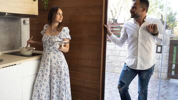 Eine Frau in einem Blumenkleid kocht in einer Küche mit Holzsärchen und Fliesenboden. Ein Mann in weißem Hemd und Jeans steht an der Tür und gestikuliert.
