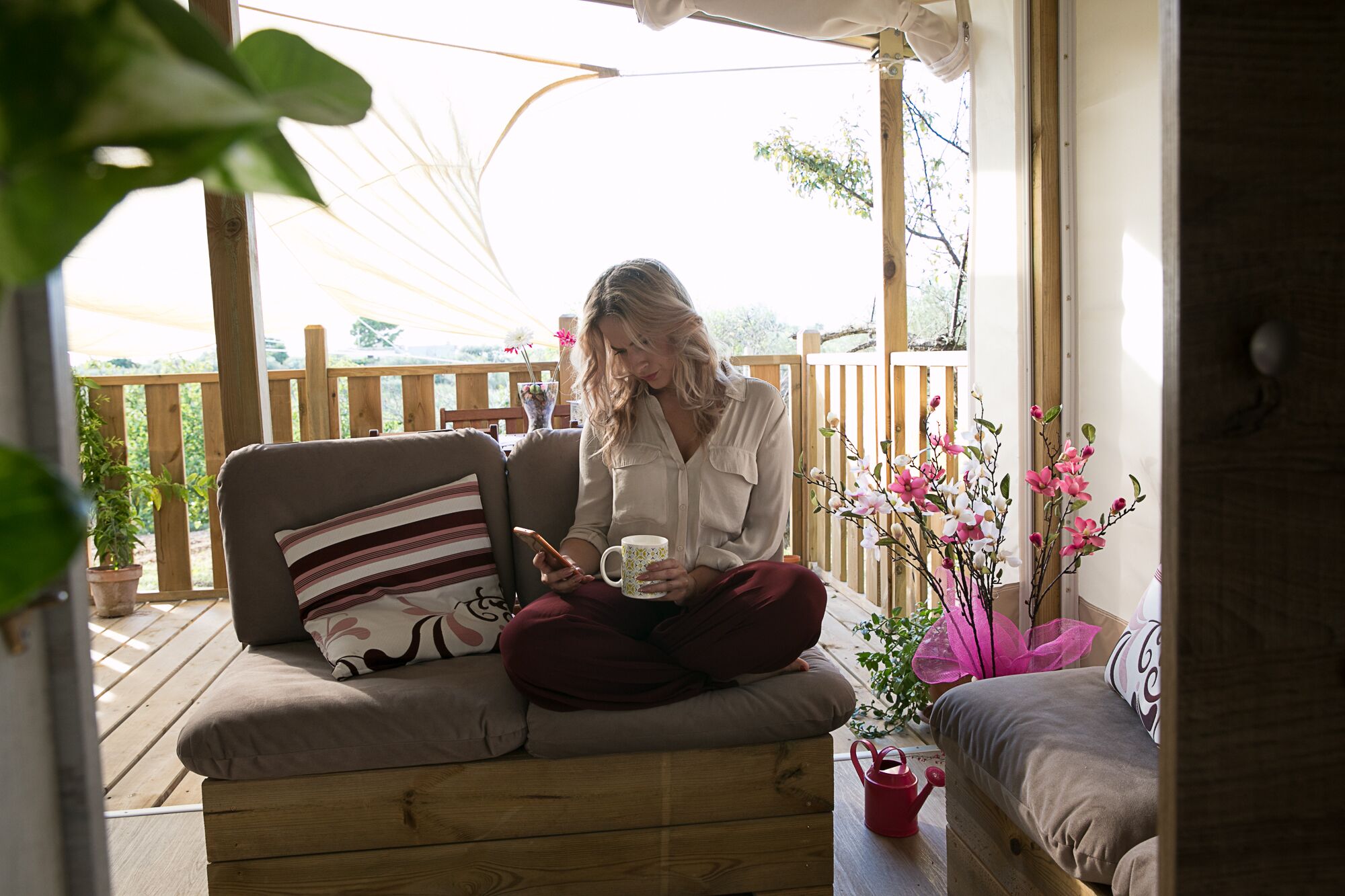 A woman sitting on a wooden bench with a cushion, holding a mug and a smartphone.