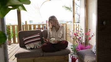 A woman sitting on a wooden bench with a cushion, holding a mug and a smartphone.