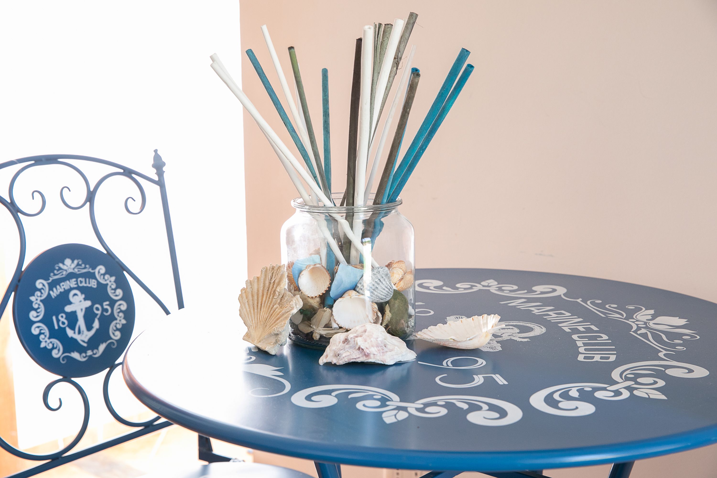 A round table with a glass jar containing seashells and reed sticks, placed on a chair with a decorative metal frame.