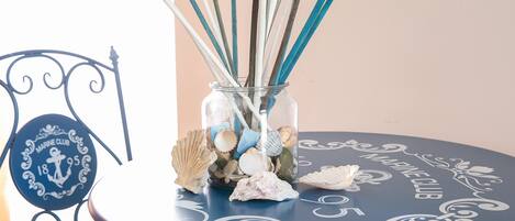 A round table with a glass jar containing seashells and reed sticks, placed on a chair with a decorative metal frame.