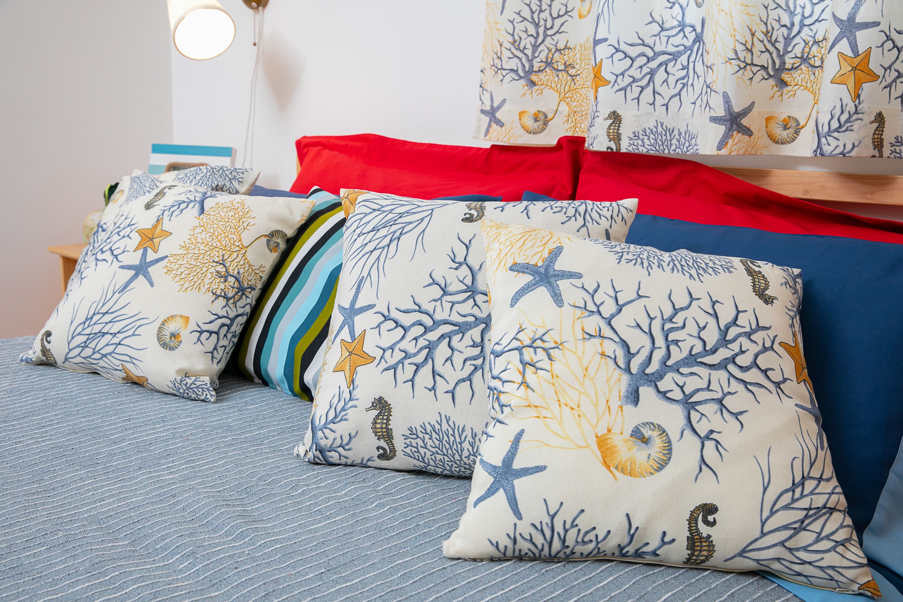 A bed with multiple pillows featuring a nautical theme, a red blanket, and a bedside table with a lamp.