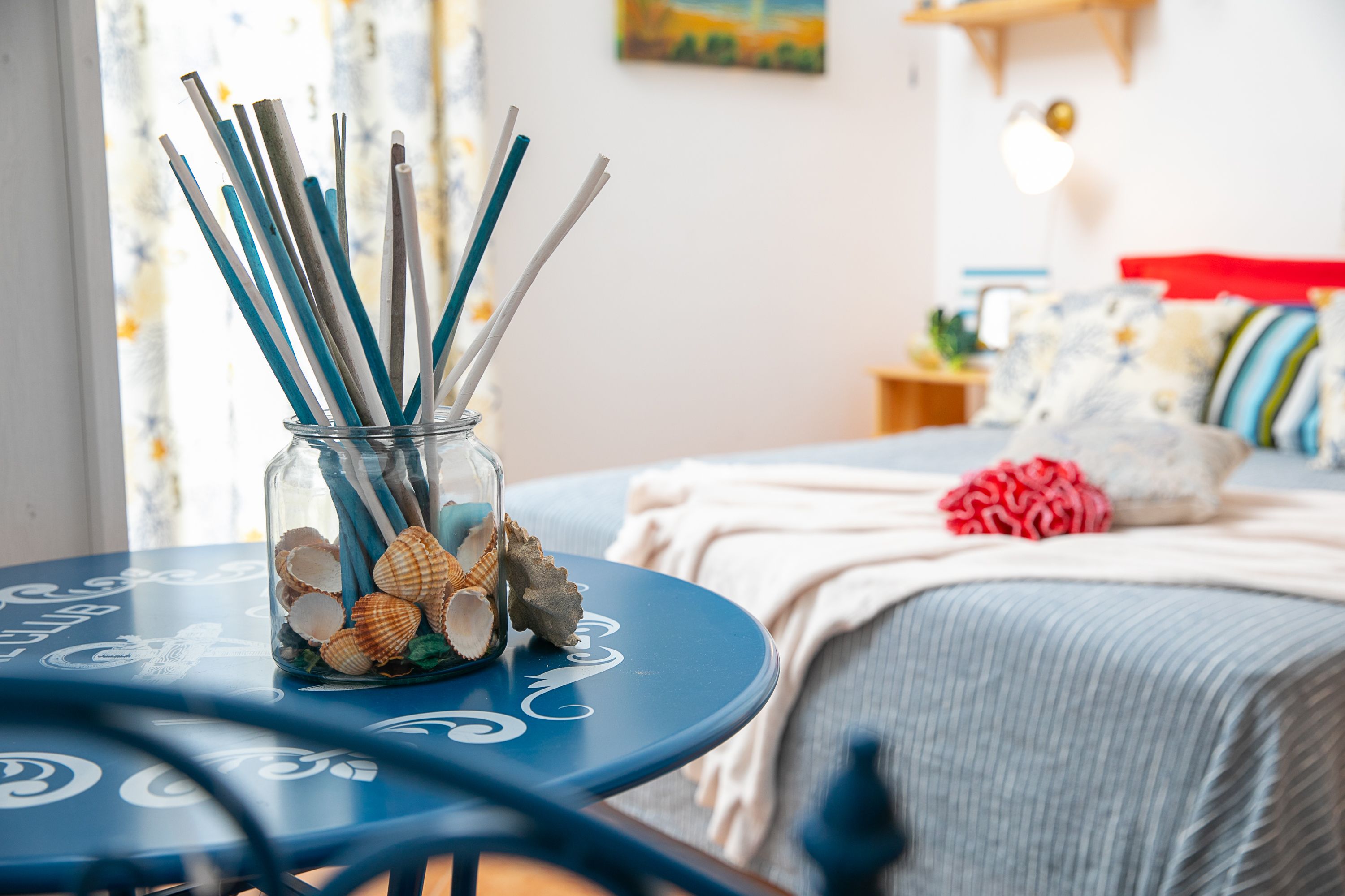 A glass jar with seashells and straws on a blue table in a room with a bed and a nightstand.