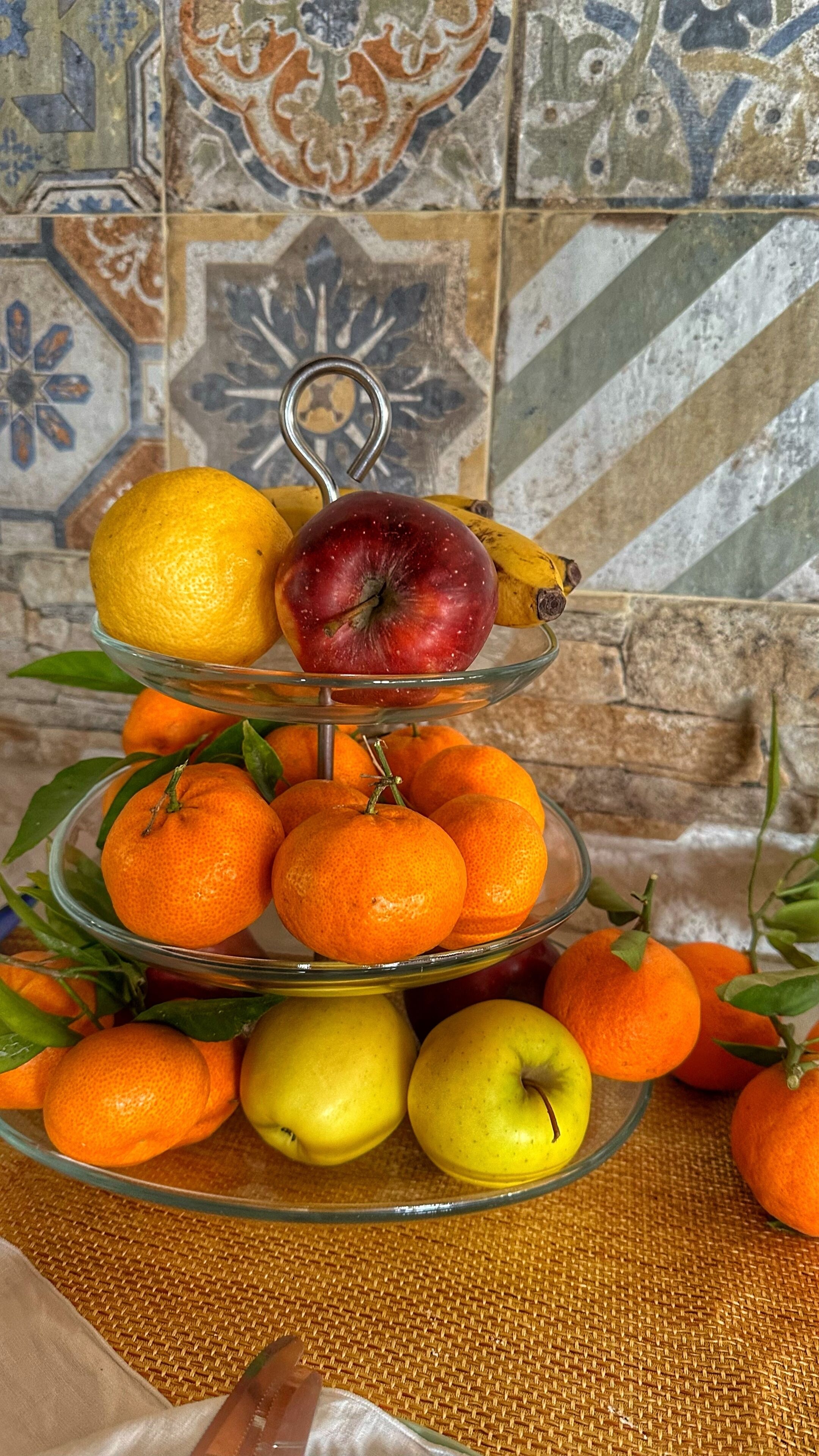 A three-tiered glass fruit stand with oranges, apples, and a banana against a tiled wall.