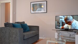 A living room with a grey sofa, a framed picture on the wall, and a television displaying a person. - 28 Scotfield (Luton)