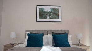 A neatly made bed with teal and white pillows, a framed abstract painting above it, and two bedside lamps on wooden nightstands. - 28 Scotfield (Luton)