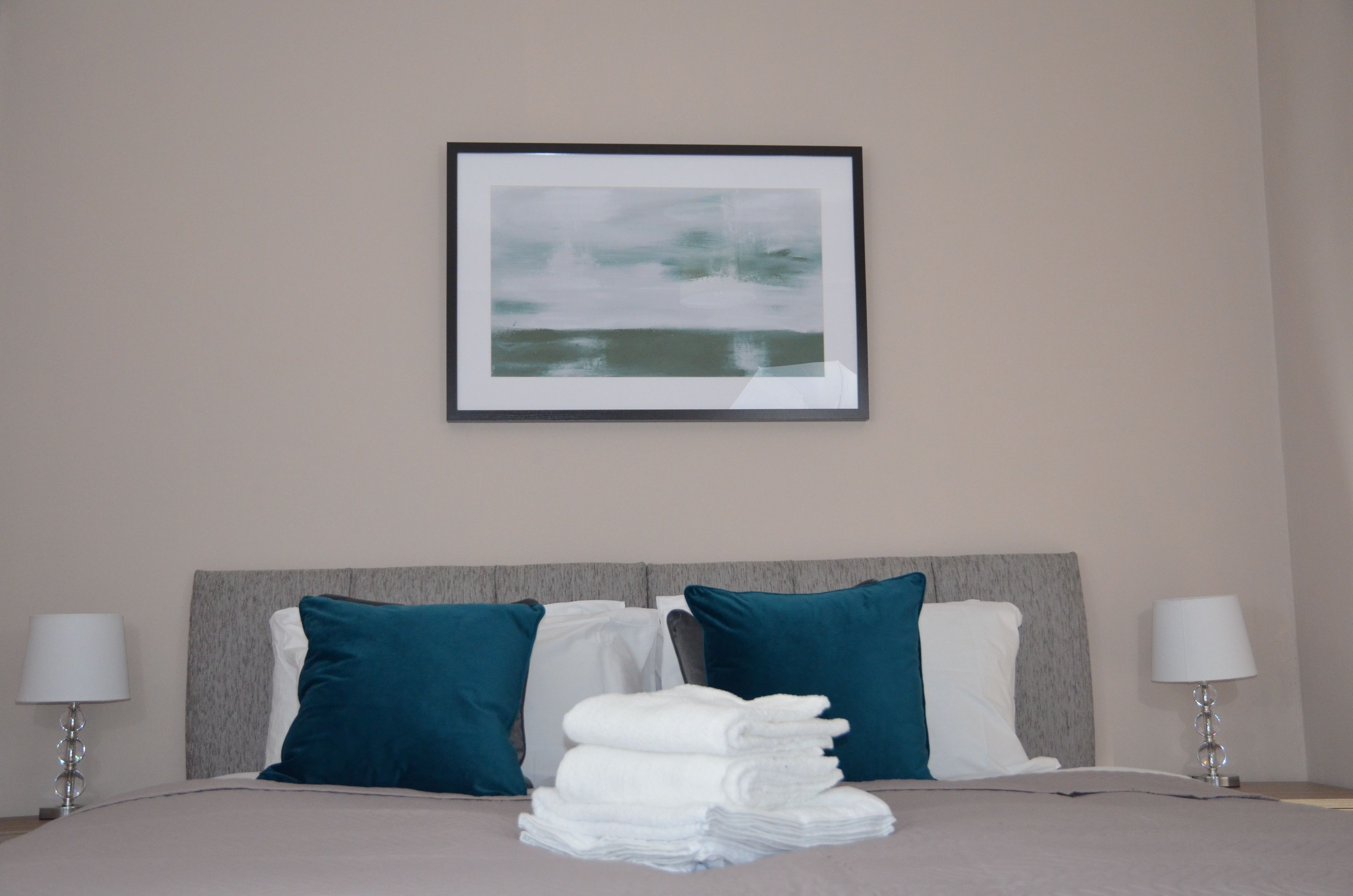 A neatly made bed with a stack of white towels, two teal pillows, and a framed abstract painting above it.