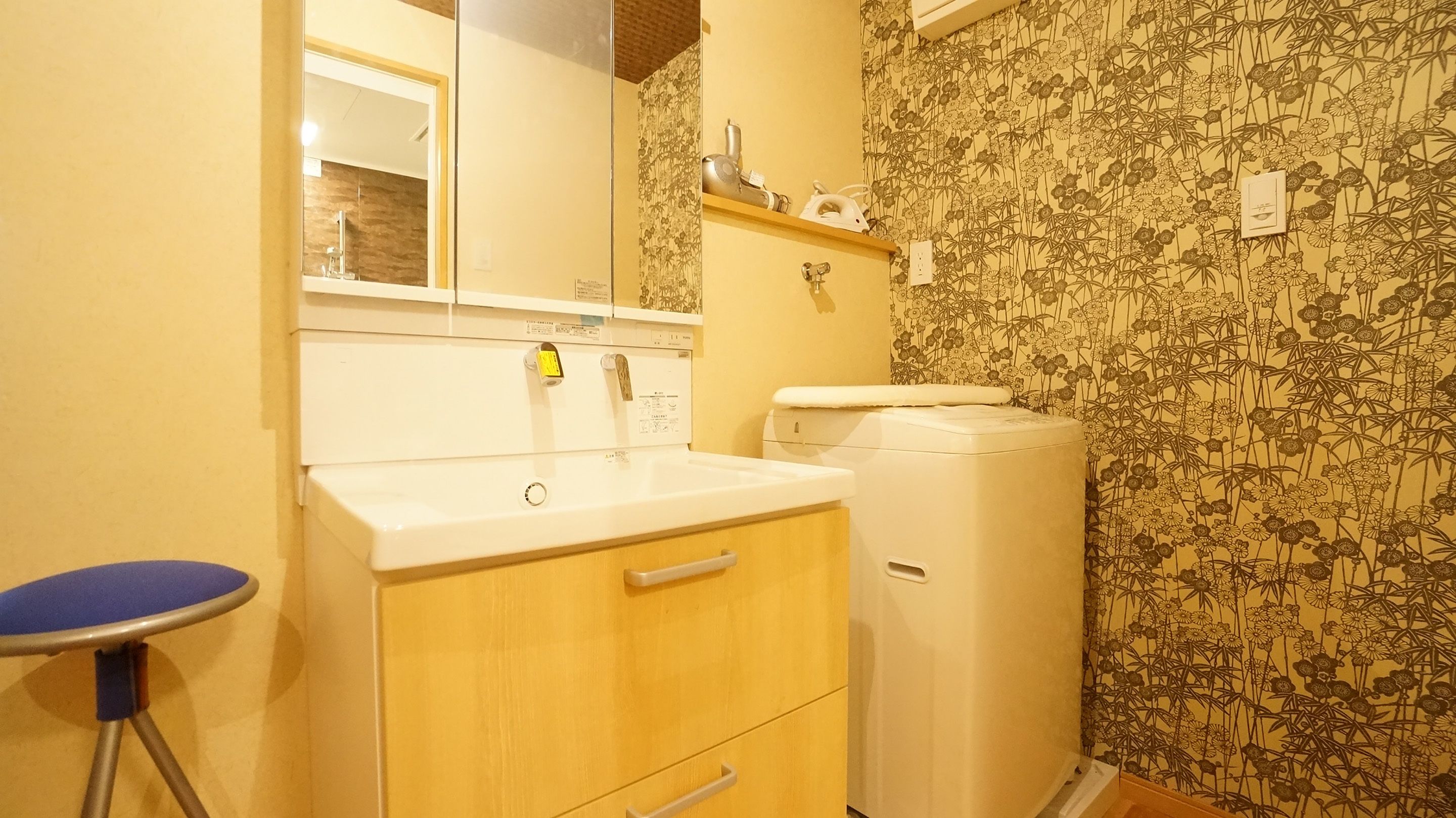 Ein Badezimmer mit Waschbecken, Spiegel und Toilette. Dazu gehören ein kleiner Hocker und ein Regal mit Pflegeprodukten.