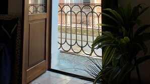 Eine geöffnete Tür, die zu einem Balkon mit schmiedeeisernem Geländer, einem Blumentopf und einem Gebäude mit rosafarbenem Mauerwerk im Hintergrund führt.