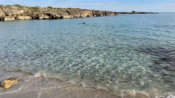 Ein Strand mit klarem Wasser, einer felsigen Küstenlinie und einer schwimmenden Person.