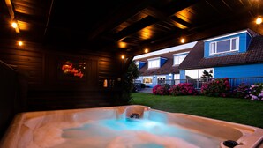 Un bain à remous sous une pergola en bois, illuminé de guirlandes lumineuses, avec un panneau néon et une maison à l’arrière-plan dont la façade est bleue.