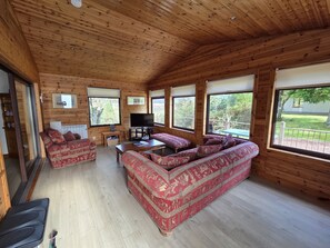 Ein Interieur einer Holzhütte mit einer Couch, einem Fernseher und großen Fenstern.