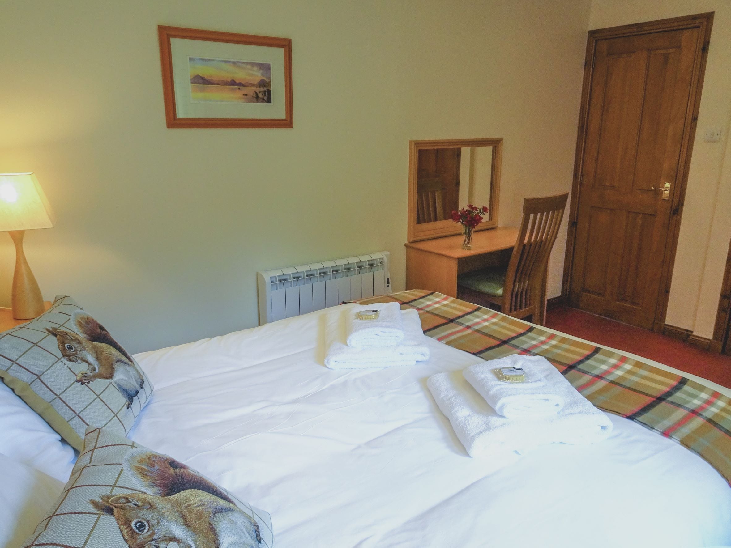 A hotel room with a bed, a bedside table with a lamp, a chair, a wooden door, and a painting on the wall.
