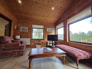 A wooden cabin interior with a sofa, a coffee table, a TV, and a window with a view of trees.