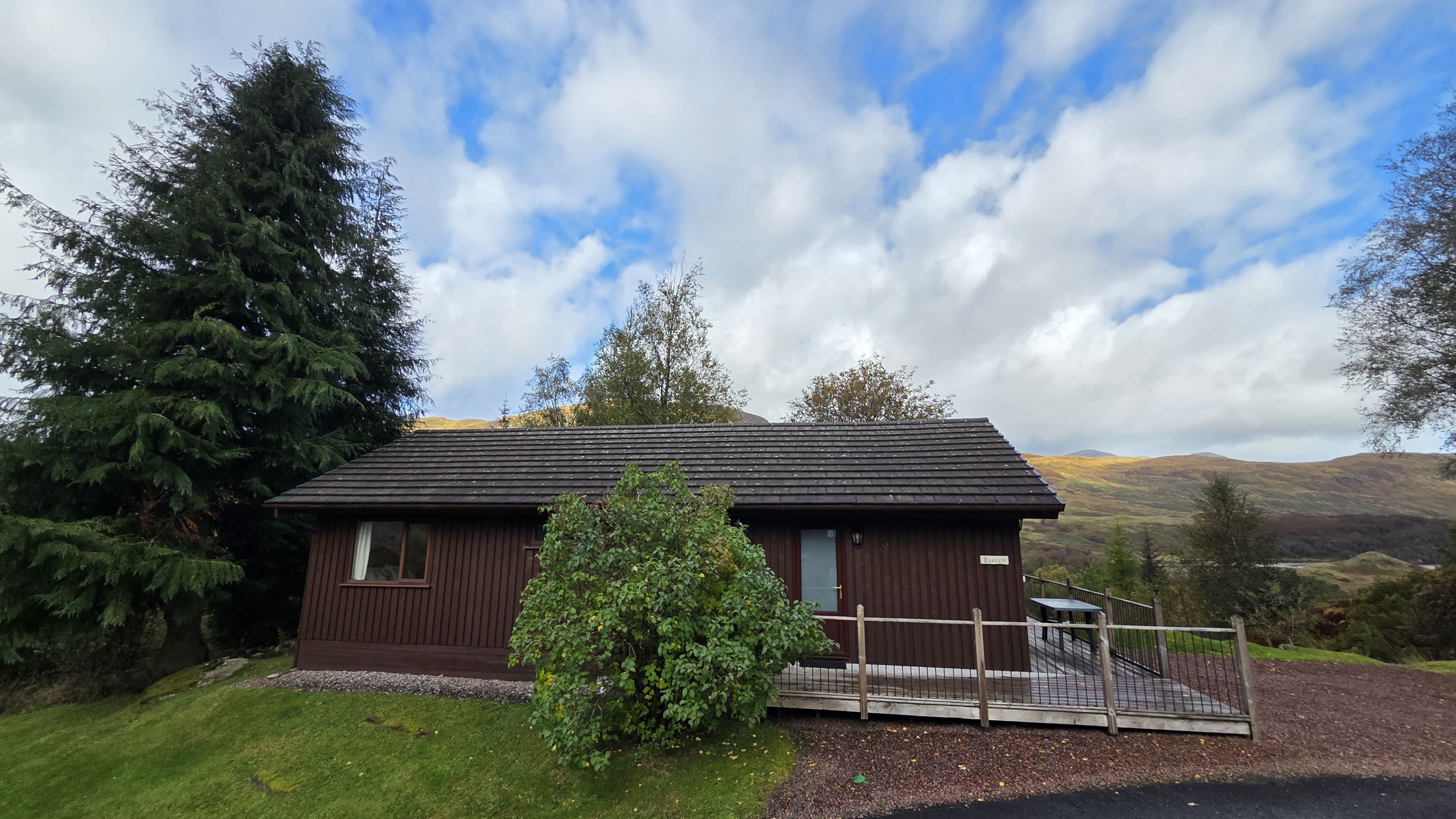 A small, single-story wooden building with a dark roof, surrounded by greenery and hills in the background.