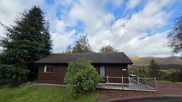 A small, single-story wooden building with a dark roof, surrounded by greenery and hills in the background.