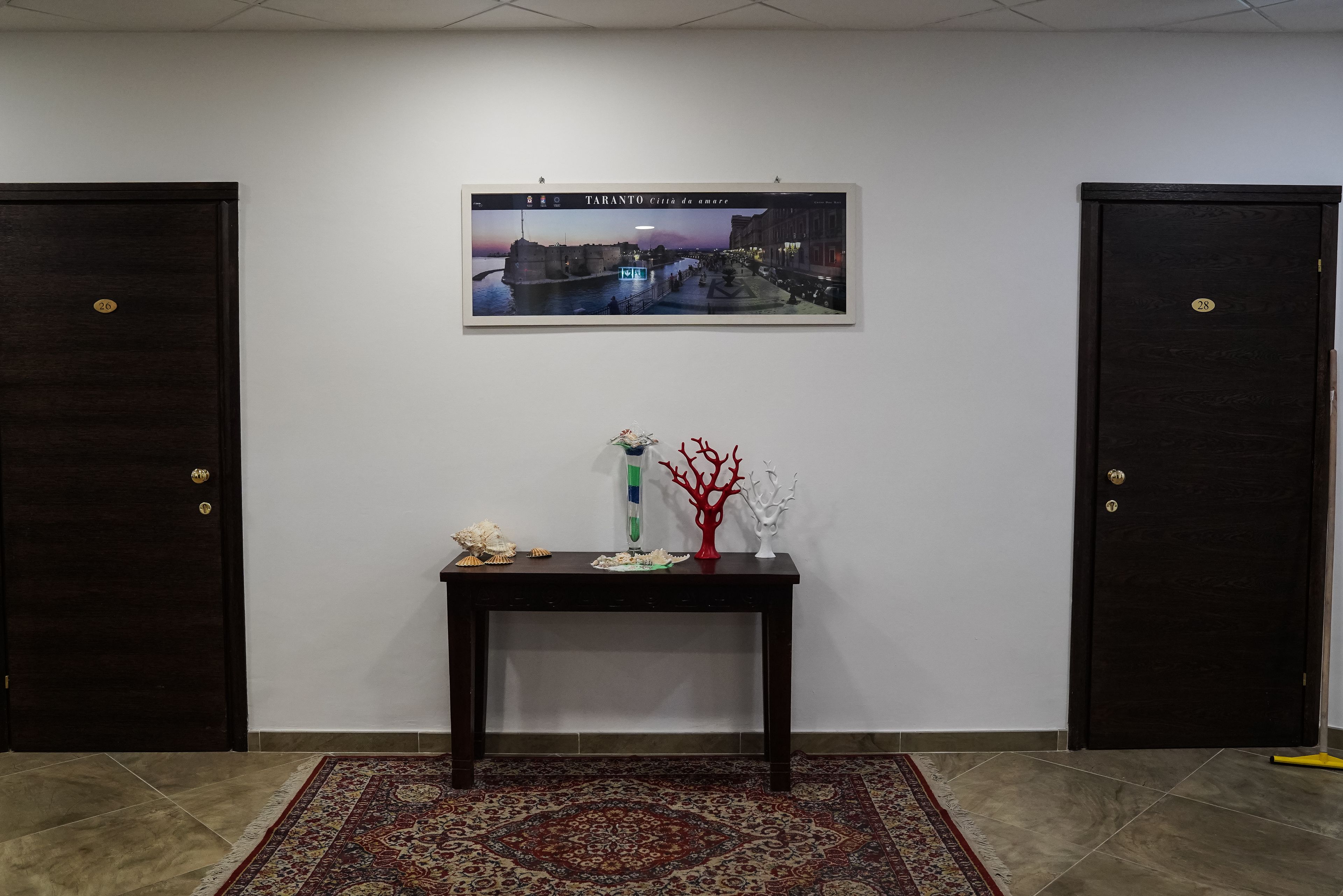 A room with two dark wooden doors, a framed picture of Taranto cityscape, a small table with decorative items, and a patterned rug.