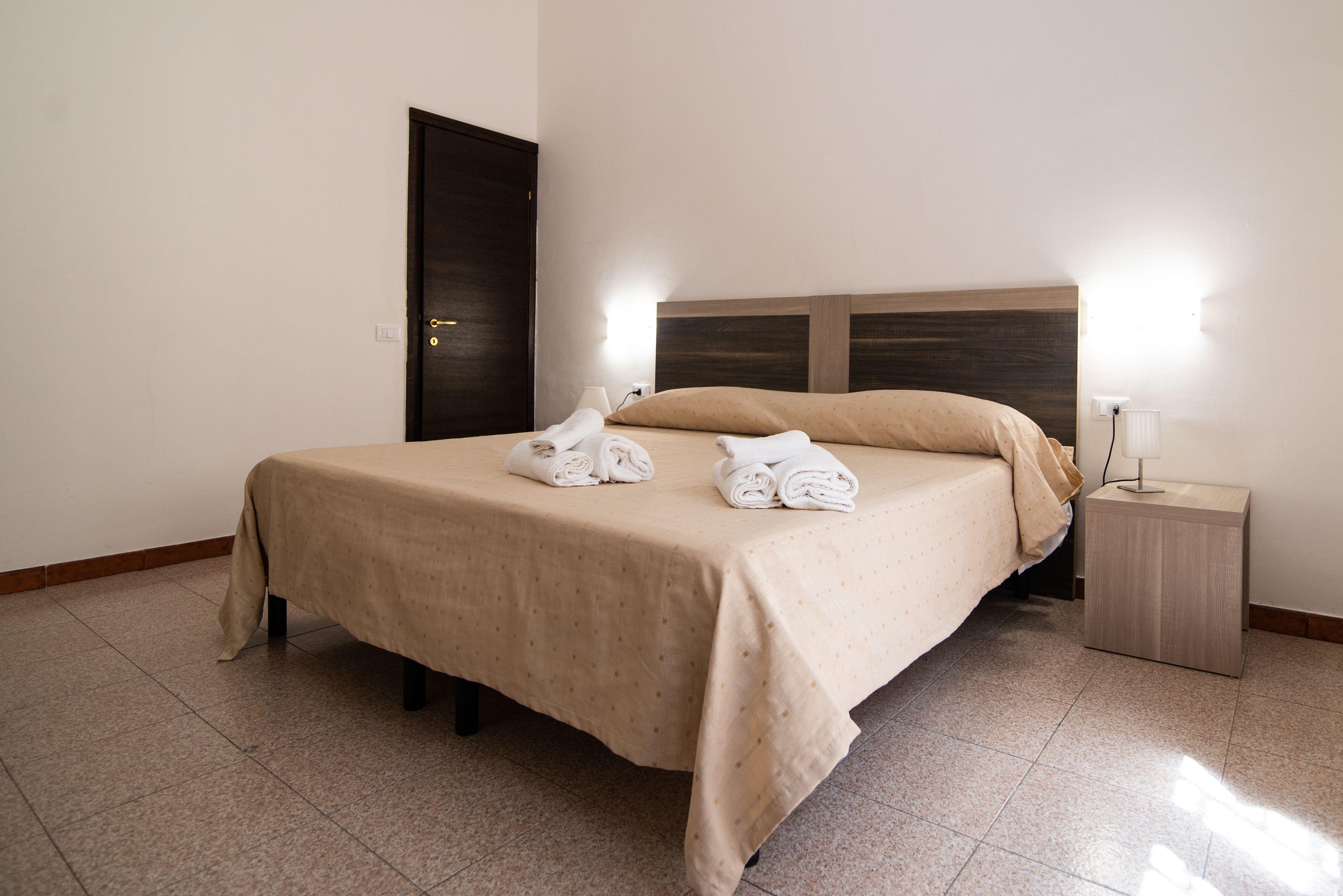 Una cama bien tendida con una colcha color beige y almohadas, una mesita de noche con una lámpara, y una puerta al fondo.