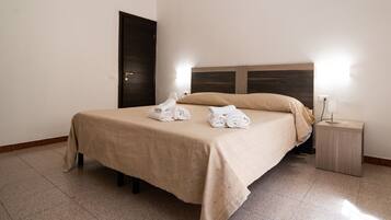 Una cama bien tendida con una colcha color beige y almohadas, una mesita de noche con una lámpara, y una puerta al fondo.