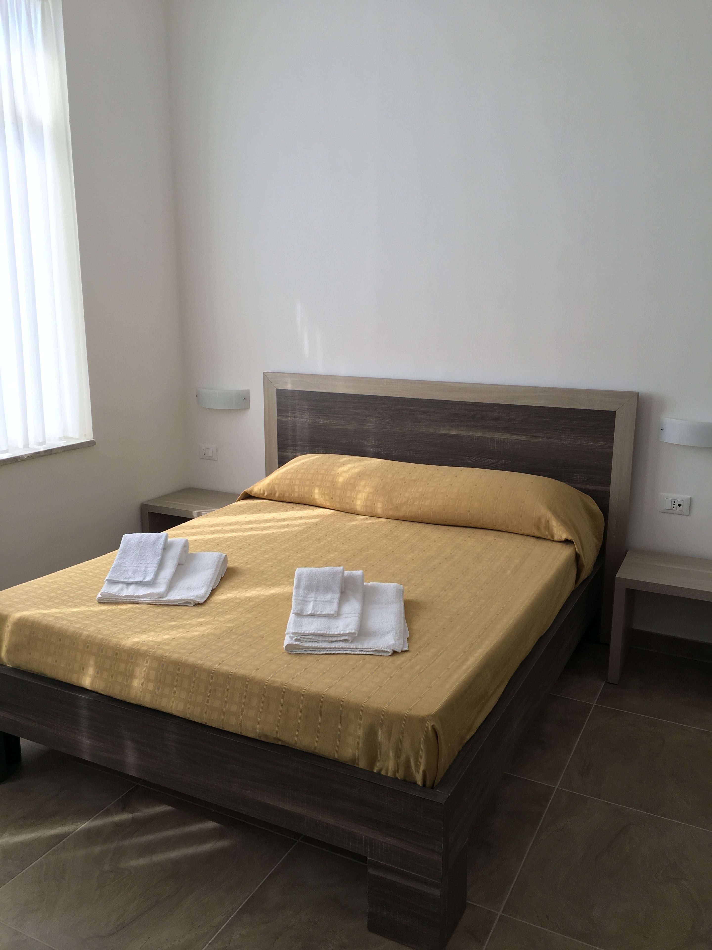 Een net opgemaakt bed met houten frame en een gele sprei, twee opgevouwen witte handdoeken op het bed, een nachtkastje met een lamp, en een raam met jaloezieën.