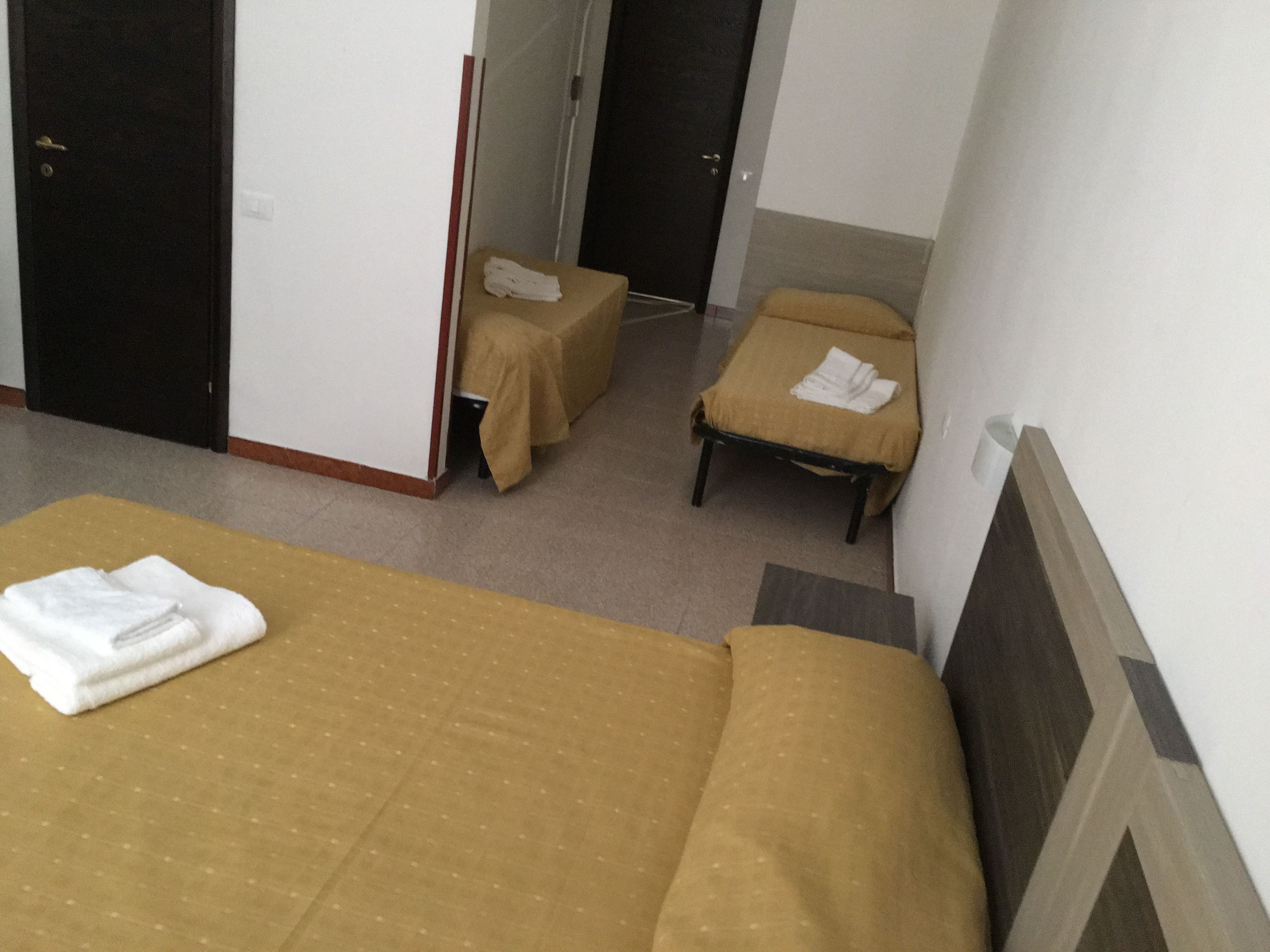 A hotel room with two single beds, each with a brown bedspread and a white towel. There are two small tables with brown tablecloths, one in front of each bed. The room has white walls and a tiled floor.